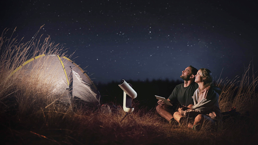 Muž se ženou sedí v noci venku a dívají se na oblohu s teleskopem Vespera.